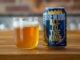 A can of Brewdog 'Ale to the King' is placed next to a clear glass filled with amber-colored beer. The can features bold, royal blue and yellow lettering with a crown design, sitting on a wooden surface with a blurred background.