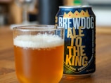 A filled glass of amber-colored beer with a frothy head sits on a wooden surface next to a can. The can features bold lettering with the words 'BREWDOG ALE TO THE KING', along with a decorative design including a crown graphic.