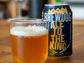 A filled glass of amber-colored beer with a frothy head sits on a wooden surface next to a can. The can features bold lettering with the words 'BREWDOG ALE TO THE KING', along with a decorative design including a crown graphic.