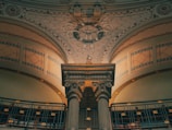 An ornate architectural interior features a classical column supporting a decorative archway. The ceiling is adorned with intricate patterns and motifs, including a roundel design. The walls are elaborately detailed with geometric and floral patterns which are highlighted by soft, warm lighting. Below, a railing with spiral ornamentation adds to the elegance of the setting.