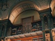 A grand, ornate interior featuring tall, decorative columns and an arched ceiling adorned with intricate patterns. A balcony lined with bookshelves surrounds the space, showcasing a collection of books. Warm lighting highlights the architectural details, creating an inviting atmosphere.