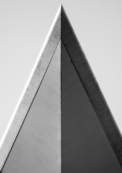 A monochrome architectural image featuring a sharp triangular structure with clean lines and contrasting surfaces. The geometric design emphasizes symmetry and minimalism.