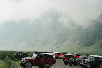 A group of riders and drivers preparing their motorcycles and 4x4 vehicles against a backdrop of towering Himalayan peaks.