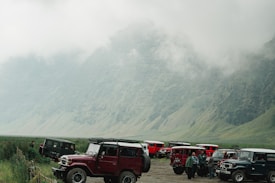 A group of off-road vehicles parked on an open landscape surrounded by mist-covered mountains, with several people standing near the vehicles. The atmosphere is serene and adventurous.