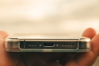 A close-up of a smartphone held horizontally by a hand, focusing on the bottom part where the charging port, speaker holes, and a transparent protective case are visible.