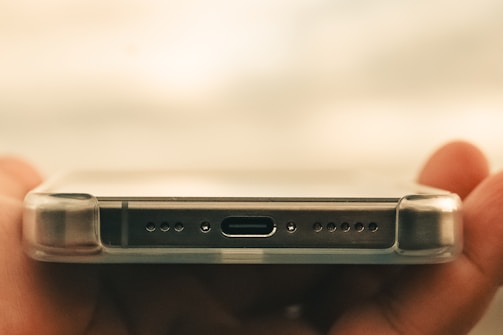 A close-up of a smartphone held horizontally by a hand, focusing on the bottom part where the charging port, speaker holes, and a transparent protective case are visible.