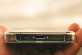 A close-up of a smartphone held horizontally by a hand, focusing on the bottom part where the charging port, speaker holes, and a transparent protective case are visible.