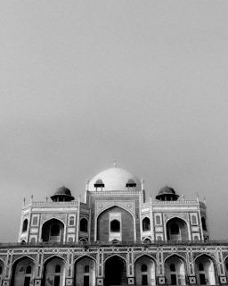 A monochrome architectural photograph features a grand historical building with an ornate facade and domed structures. The intricate detailing and symmetry of arches highlight the building's majestic design.