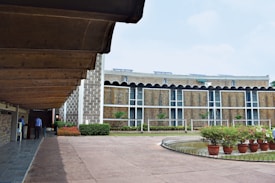 A modern architectural building with a textured facade featuring vertical patterns, surrounded by a well-maintained courtyard and garden area. There are potted plants neatly placed around a circular water feature on the right side, while two individuals are seen walking under a covered walkway on the left.