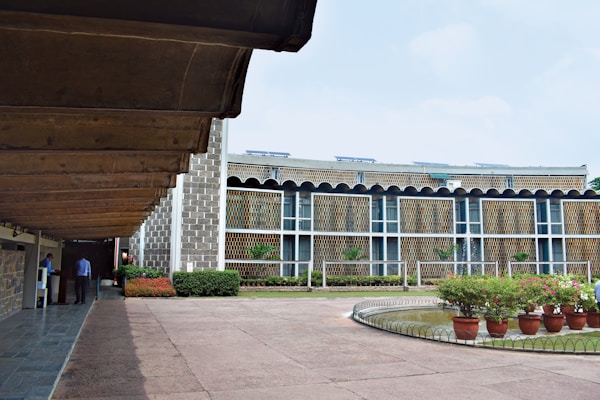 A modern architectural building with a textured facade featuring vertical patterns, surrounded by a well-maintained courtyard and garden area. There are potted plants neatly placed around a circular water feature on the right side, while two individuals are seen walking under a covered walkway on the left.