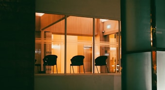 A dimly lit room is visible through large windows, showcasing a series of empty black chairs arranged against a warm yellow-orange backdrop. The lighting creates a cozy atmosphere, with shadows cast from the chairs adding depth. The room appears modern, with clean lines and a hint of a door labeled with an exit sign.