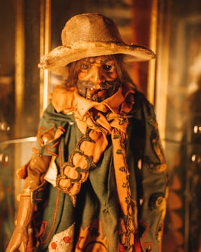 A detailed, rustic puppet with a rugged appearance adorned in an intricately decorated outfit featuring green and orange hues. The puppet has a wide-brimmed hat and a weathered face with exaggerated features, giving it an aged, artisanal look.