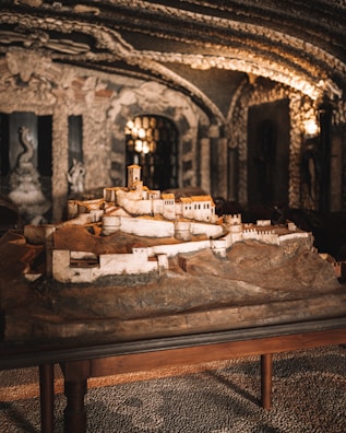 A detailed miniature model of an ancient fortress, constructed with earthy tones and positioned on a wooden table. The background features ornately decorated walls and ceilings featuring baroque-style sculptures and arches with ambient lighting, setting an antiquated and artistic atmosphere.