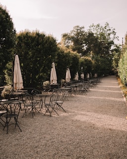 An outdoor patio area lined with black metal chairs and tables. Beige closed umbrellas are evenly spaced along the edge of the space. Tall lush hedges form a natural wall on either side, creating an intimate path along the pebbled ground.