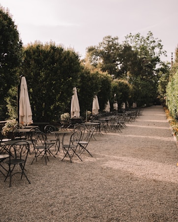 An outdoor patio area lined with black metal chairs and tables. Beige closed umbrellas are evenly spaced along the edge of the space. Tall lush hedges form a natural wall on either side, creating an intimate path along the pebbled ground.
