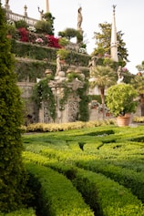 A beautifully manicured garden featuring dense green hedges arranged in intricate patterns. Statues of various figures stand atop stone pillars, surrounded by vibrant flowers and lush greenery. Stonework and architectural details add to the classical and enchanting atmosphere.