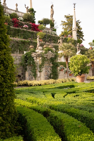 A beautifully manicured garden featuring dense green hedges arranged in intricate patterns. Statues of various figures stand atop stone pillars, surrounded by vibrant flowers and lush greenery. Stonework and architectural details add to the classical and enchanting atmosphere.