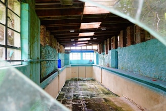 An abandoned indoor pool room with weathered brick and turquoise painted walls. The ceiling is made of exposed wooden beams, and the floor of the empty pool shows dark, wet patches. Light streams in through large windows at one end, with broken glass visible in the foreground.