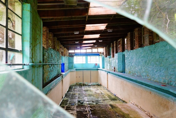An abandoned indoor pool room with weathered brick and turquoise painted walls. The ceiling is made of exposed wooden beams, and the floor of the empty pool shows dark, wet patches. Light streams in through large windows at one end, with broken glass visible in the foreground.