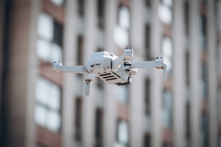 A small white drone hovers in the air with blurred high-rise apartment buildings in the background. The drone's detailed features, including its four rotors and camera, are in clear focus.