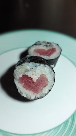 Two sushi rolls, consisting of rice and a piece of pink fish at the center, are wrapped in nori seaweed. They sit on a white plate with a green rim, and the lighting creates a slight shadow around them.