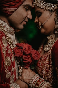 A couple dressed in traditional, richly embroidered attire is holding a bouquet of red roses. The bride is adorned with ornate jewelry, including a nose ring and forehead jewelry, and intricate henna designs can be seen on her hands. They are intimately close, with expressions of joy and affection.