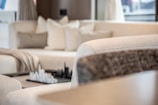 A modern living room featuring a plush cream-colored sectional sofa with several cushions. A decorative chess set is placed on a small table in the background, adding a sophisticated touch. Soft natural light filters through the space, creating a serene atmosphere.