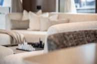 A modern living room featuring a plush cream-colored sectional sofa with several cushions. A decorative chess set is placed on a small table in the background, adding a sophisticated touch. Soft natural light filters through the space, creating a serene atmosphere.
