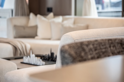A modern living room featuring a plush cream-colored sectional sofa with several cushions. A decorative chess set is placed on a small table in the background, adding a sophisticated touch. Soft natural light filters through the space, creating a serene atmosphere.