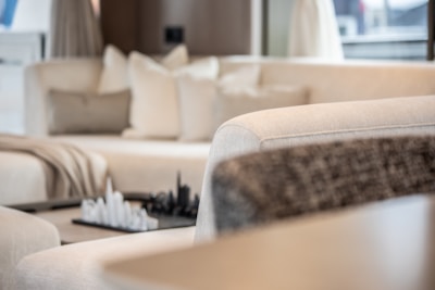 A modern living room featuring a plush cream-colored sectional sofa with several cushions. A decorative chess set is placed on a small table in the background, adding a sophisticated touch. Soft natural light filters through the space, creating a serene atmosphere.