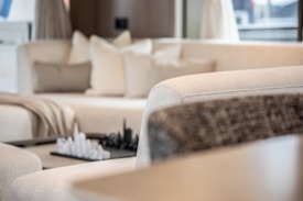 A modern living room featuring a plush cream-colored sectional sofa with several cushions. A decorative chess set is placed on a small table in the background, adding a sophisticated touch. Soft natural light filters through the space, creating a serene atmosphere.