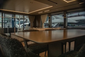 A luxurious interior of a yacht featuring a large dining table and upholstered chairs. The large windows provide a view of another yacht parked nearby, with curtains partially drawn. The atmosphere is elegant with soft lighting from recessed ceiling lights.