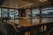 A luxurious interior of a yacht featuring a large dining table and upholstered chairs. The large windows provide a view of another yacht parked nearby, with curtains partially drawn. The atmosphere is elegant with soft lighting from recessed ceiling lights.