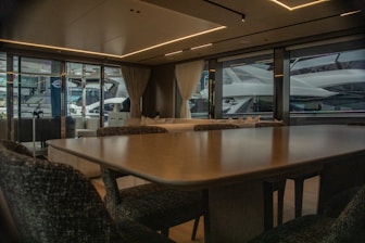 A luxurious interior of a yacht featuring a large dining table and upholstered chairs. The large windows provide a view of another yacht parked nearby, with curtains partially drawn. The atmosphere is elegant with soft lighting from recessed ceiling lights.
