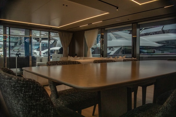 A luxurious interior of a yacht featuring a large dining table and upholstered chairs. The large windows provide a view of another yacht parked nearby, with curtains partially drawn. The atmosphere is elegant with soft lighting from recessed ceiling lights.