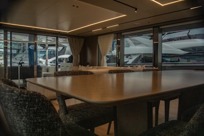 A luxurious interior of a yacht featuring a large dining table and upholstered chairs. The large windows provide a view of another yacht parked nearby, with curtains partially drawn. The atmosphere is elegant with soft lighting from recessed ceiling lights.