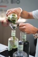 A person is pouring green liquid from a glass jar into a funnel placed on top of a bottle labeled 'Anaë.' The scene is focused on the hands and the bottles, with a watch visible on the person's wrist. There are multiple bottles in the foreground, and the setting appears to be indoors, possibly in a laboratory or kitchen.