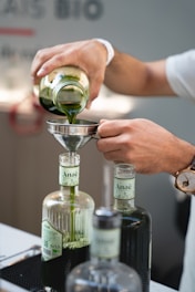 A person is pouring green liquid from a glass jar into a funnel placed on top of a bottle labeled 'Anaë.' The scene is focused on the hands and the bottles, with a watch visible on the person's wrist. There are multiple bottles in the foreground, and the setting appears to be indoors, possibly in a laboratory or kitchen.