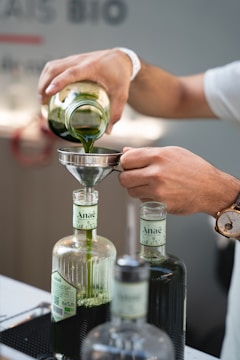 A person is pouring green liquid from a glass jar into a funnel placed on top of a bottle labeled 'Anaë.' The scene is focused on the hands and the bottles, with a watch visible on the person's wrist. There are multiple bottles in the foreground, and the setting appears to be indoors, possibly in a laboratory or kitchen.