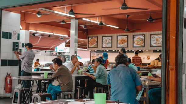A bustling indoor food court features several people seated at round tables, engaged in eating and conversation. The decor includes ceiling fans, framed food pictures on the wall, and bright lighting. A person stands near a wall-mounted device, possibly a ticket machine, while others at the tables appear to be dining. The setting feels communal and lively.