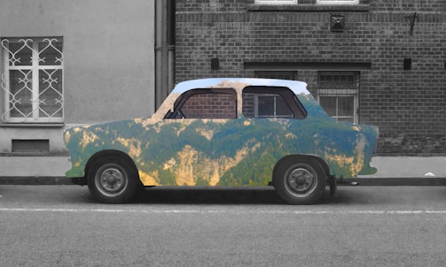 A vintage car is parked on a street with a unique artistic design. The car's body features a colorful landscape pattern resembling mountainous terrain, contrasting with the grayscale environment. The background includes a brick building and a barred window.
