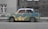 A vintage car is parked on a street with a unique artistic design. The car's body features a colorful landscape pattern resembling mountainous terrain, contrasting with the grayscale environment. The background includes a brick building and a barred window.
