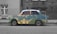 A vintage car is parked on a street with a unique artistic design. The car's body features a colorful landscape pattern resembling mountainous terrain, contrasting with the grayscale environment. The background includes a brick building and a barred window.