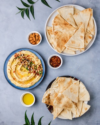 A flat lay composition featuring a bowl of hummus garnished with herbs and spices on a blue plate, surrounded by two plates of pita bread cut into triangles. There are small bowls containing almonds, chili flakes, and olive oil. The background is a light gray surface with a few green leaves adding a touch of color.