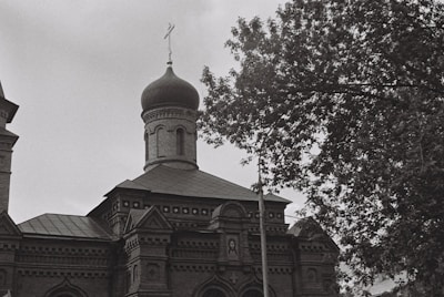 A black and white photograph of an ornate building with a prominent onion-shaped dome typical of Russian architecture. The building features intricate brickwork and decorative elements. A tree with lush foliage partially obscures the structure on the right side.