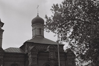 A black and white photograph of an ornate building with a prominent onion-shaped dome typical of Russian architecture. The building features intricate brickwork and decorative elements. A tree with lush foliage partially obscures the structure on the right side.