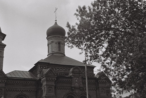 A black and white photograph of an ornate building with a prominent onion-shaped dome typical of Russian architecture. The building features intricate brickwork and decorative elements. A tree with lush foliage partially obscures the structure on the right side.