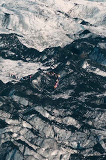 A group of people, wearing brightly colored clothing and gear, traverse across a vast, rugged terrain dominated by contrasting dark and light surfaces. The landscape appears icy and cold, with jagged patterns covering the ground.