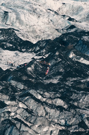 A group of people, wearing brightly colored clothing and gear, traverse across a vast, rugged terrain dominated by contrasting dark and light surfaces. The landscape appears icy and cold, with jagged patterns covering the ground.