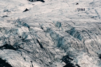 A vast, rugged glacier landscape with noticeable crevasses, showing a group of people standing in the distance on the glacier surface. The ice has a textured, layered appearance with dark ridges running through it.